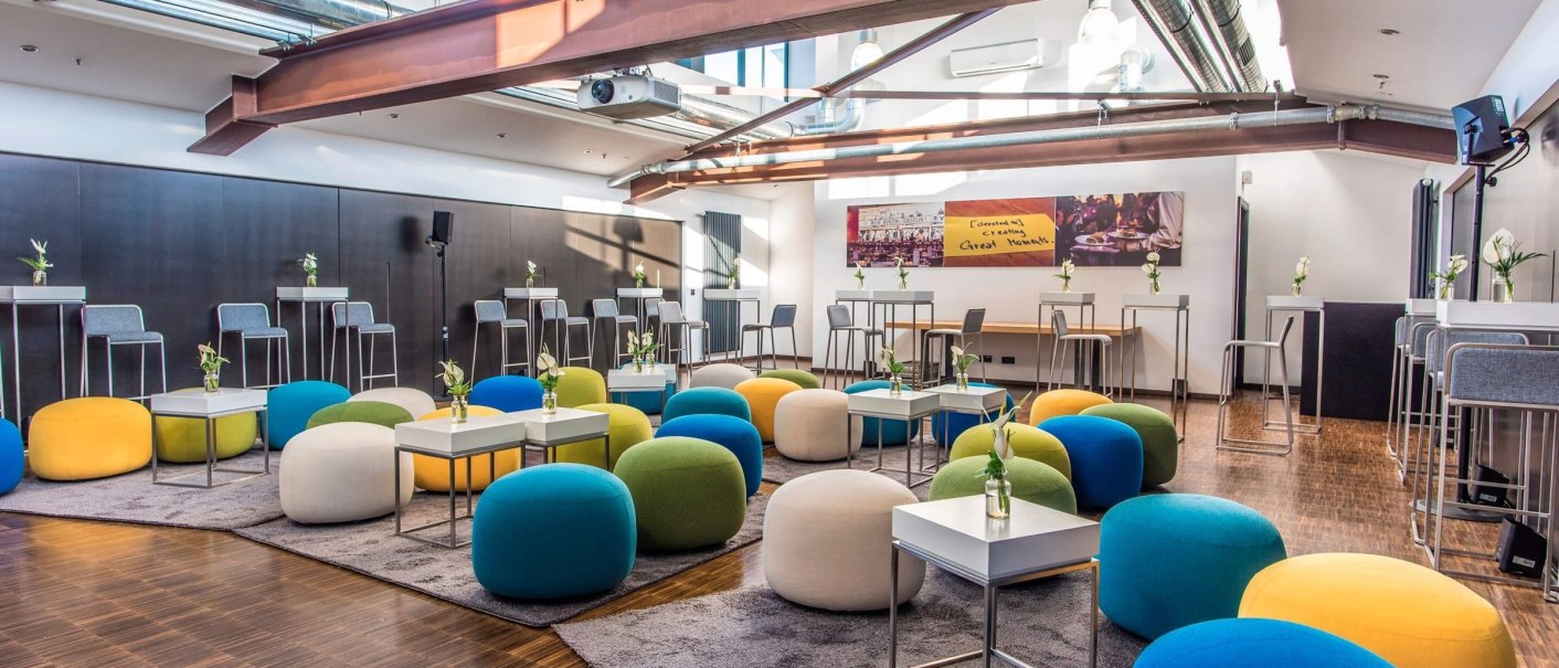 Modern seminar room with colorful seat cushions, small tables and high chairs. Large windows and exposed ceiling beams create an open ambience., © rauschenbergercatering Modern seminar room with colorful seat cushions, small tables and high chairs. Large windows and exposed ceiling beams create an open ambience., © rauschenbergercatering