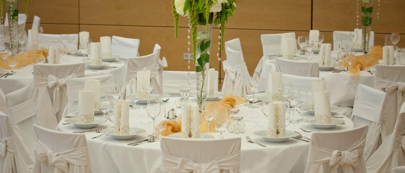 Elegante Tischdekoration mit weißen Stuhlhussen, Kerzen und Blumenarrangements in einem festlichen Saal., © Linda Helbing