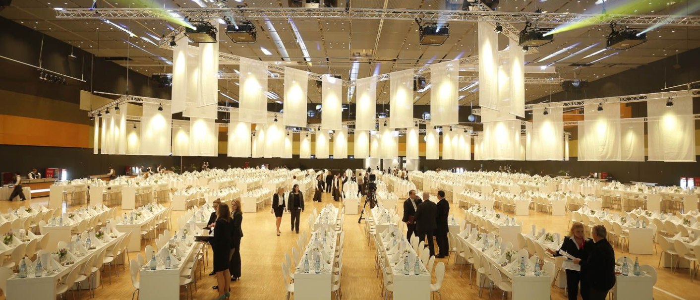 Ein großer Kongresssaal mit langen, gedeckten Tischen. Menschen in formeller Kleidung bereiten sich auf eine Veranstaltung vor., © Messe Stuttgart Ein großer Kongresssaal mit langen, gedeckten Tischen. Menschen in formeller Kleidung bereiten sich auf eine Veranstaltung vor., © Messe Stuttgart
