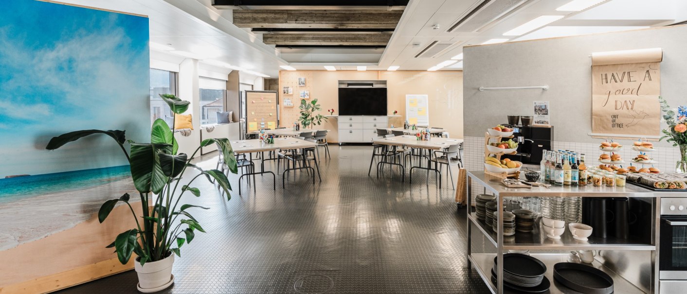 Moderner Raum mit Tischen, Stühlen, Snack-Bar und großem Bildschirm. Pflanzen und Strandbild als Dekoration., © OutOfOffice GmbH Moderner Raum mit Tischen, Stühlen, Snack-Bar und großem Bildschirm. Pflanzen und Strandbild als Dekoration., © OutOfOffice GmbH