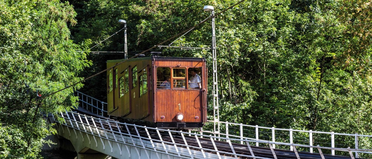 Eine historische Seilbahn mit Holzverkleidung fährt durch einen dichten Wald auf einer Schiene., © TBMW, Gregor Lengler