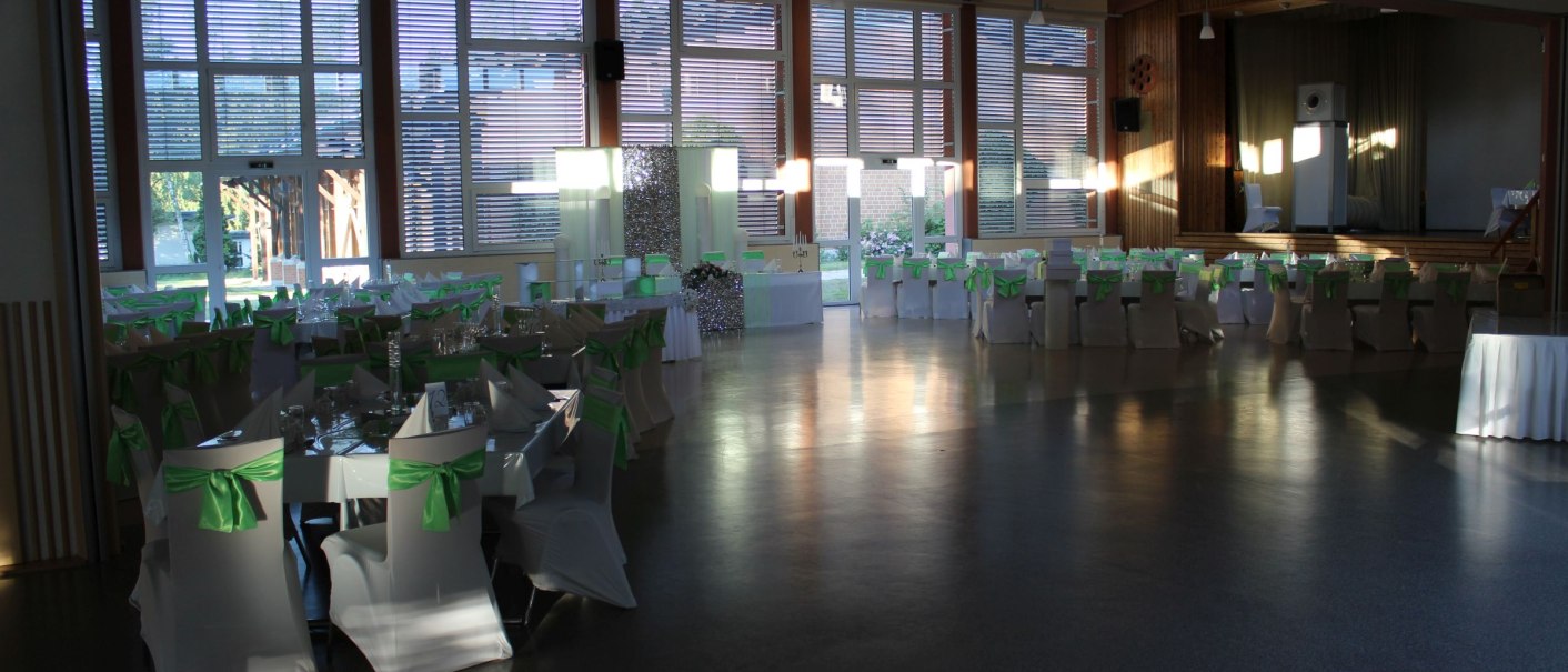 Festlich dekorierte Halle mit gedeckten Tischen und St&uuml;hlen, die mit gr&uuml;nen Schleifen verziert sind. Gro&szlig;e Fenster lassen Licht herein., &copy; Gemeinde Alth&uuml;tte