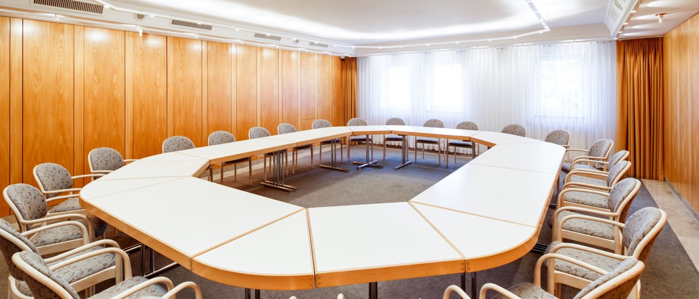 Konferenzraum mit U-förmigem Tisch und gepolsterten Stühlen, helle Holzvertäfelung, weiße Vorhänge, Deckenbeleuchtung., © Tobias Fröhner Photography