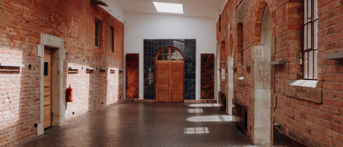 Ein langer Korridor mit roten Backsteinwänden, Oberlichtern und einer Holztür am Ende. Sonnenlicht fällt durch die Fenster auf den Boden., © Exometa Römerkastell Ein langer Korridor mit roten Backsteinwänden, Oberlichtern und einer Holztür am Ende. Sonnenlicht fällt durch die Fenster auf den Boden., © Exometa Römerkastell