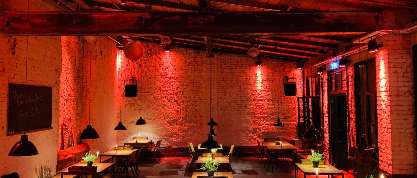 Small room with brick walls, illuminated in red. Tables and chairs are decorated with plants and candles. Cozy atmosphere., © Wagenhallen Stuttgart GmbH & Co. KG