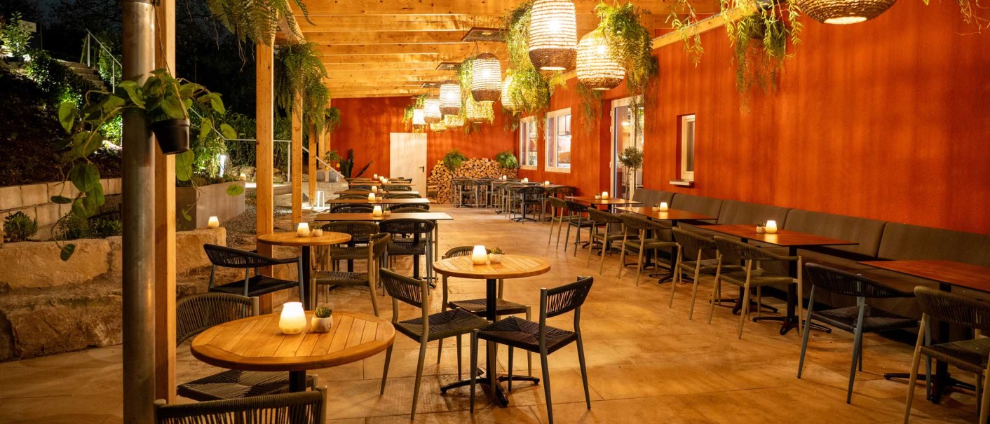 Cozy outdoor area with wooden tables, chairs and hanging plants. Warm lighting and red wall create an inviting atmosphere., © SatzDrei