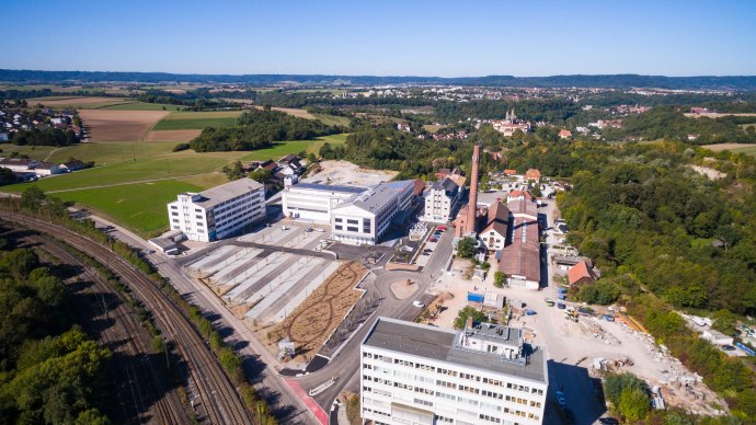 Luftaufnahme einer Fabrikanlage mit mehreren Gebäuden und Schornstein, umgeben von Feldern und Wäldern. Im Hintergrund sind Hügel und eine Stadt zu sehen., © Stefan Baraniecki