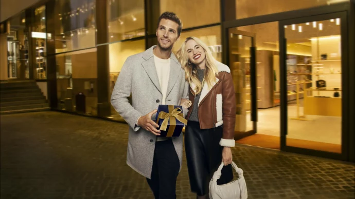 L&auml;chelndes Paar vor einem beleuchteten Gesch&auml;ft, der Mann mit Geschenkbox, die Frau mit Handtasche. Weihnachtlich und elegant., &copy; OUTLETCITY AG