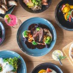 Ein Tisch mit verschiedenen Gerichten: Fleisch mit Gemüse, Salate, Burger mit Pommes, Brot und Desserts. Getränke sind ebenfalls zu sehen., © SatzDrei