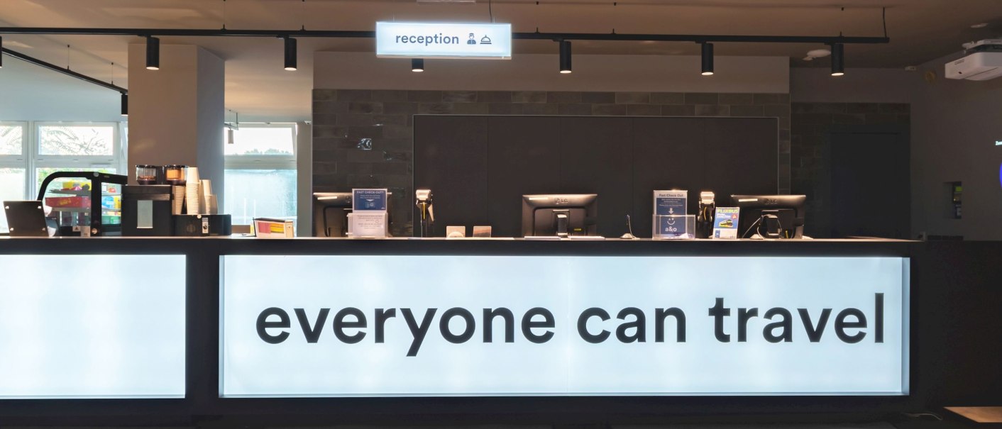 Moderne Rezeption im a&o Stuttgart City mit beleuchtetem Tresen und dem Schriftzug 'everyone can travel'., © a&o Hostels