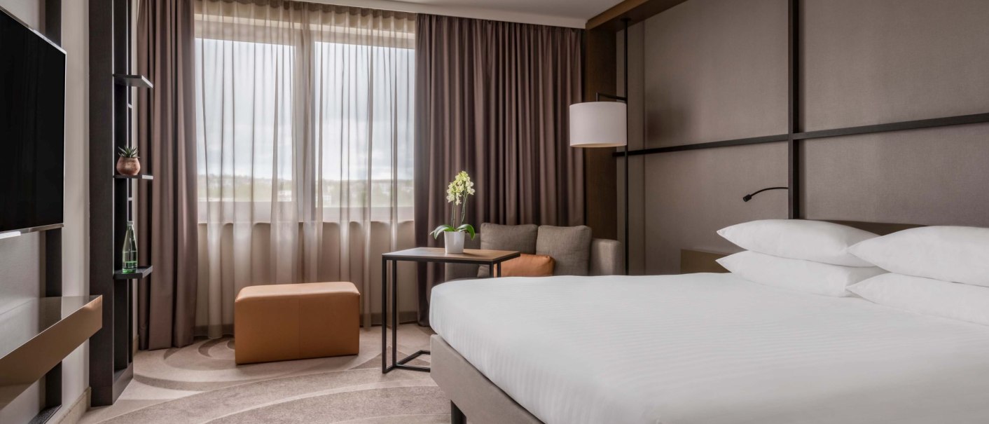 Modernes Hotelzimmer mit großem Bett, Sofa, Tisch und Fenster mit Vorhängen. Dezente Farben und stilvolle Einrichtung., © Stuttgart Marriott Hotel Sindelfingen, Kilian Blees