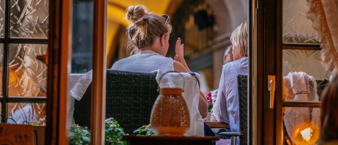 Zwei Personen sitzen in einem Café, von außen durch ein Fenster gesehen. Eine Person hat einen Dutt, die Atmosphäre wirkt gemütlich und einladend., © Thomas Niedermüller
