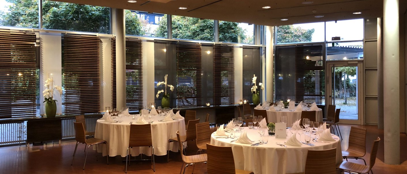 Elegant room with round, laid tables, wooden chairs and large windows. Subtle lighting and plants create a stylish atmosphere., © Filharmonie_gf