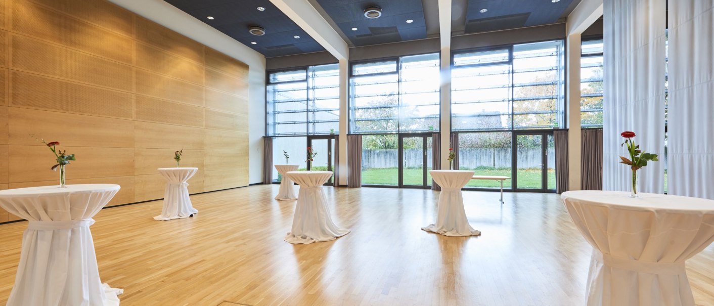 Large, bright room with wooden floor, bar tables with white tablecloths and flowers. Large window front with a view of the greenery., &copy; bebopmedia lukas breusch