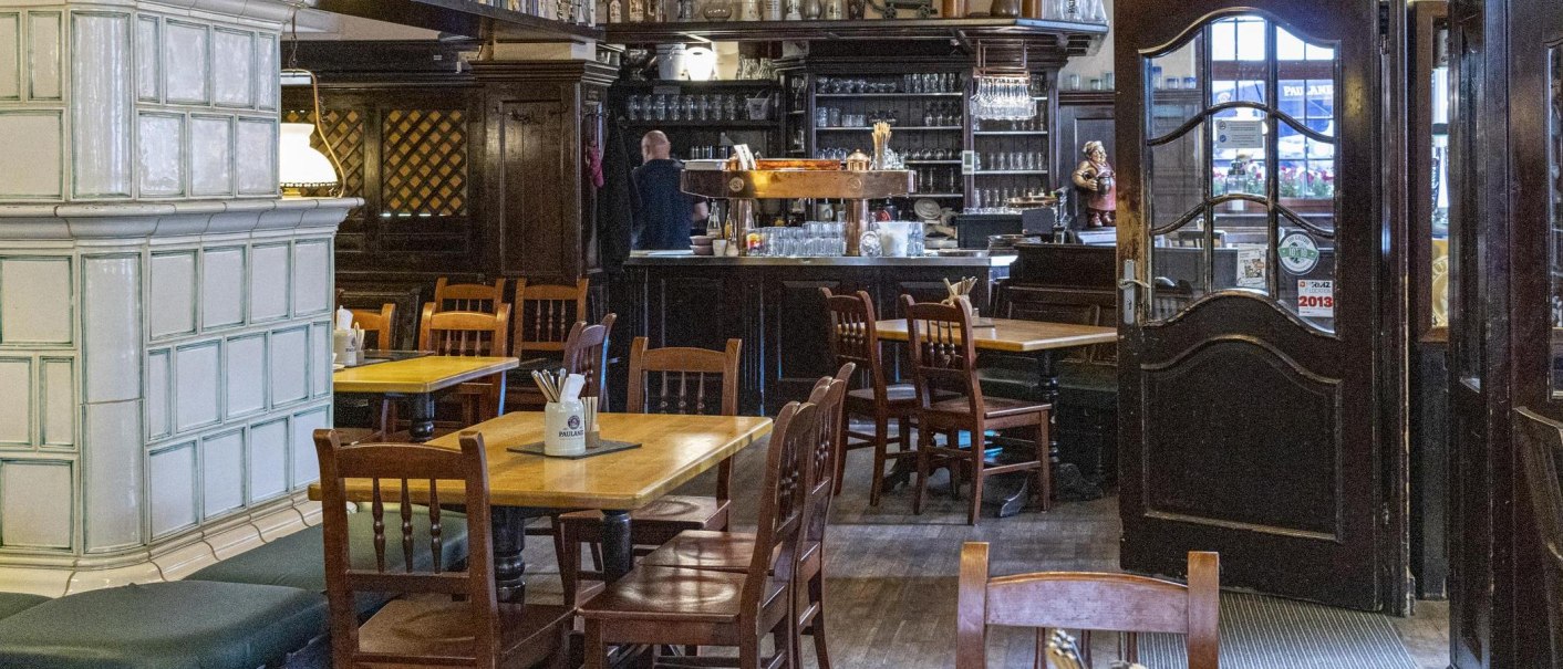 Cozy interior of a restaurant with wooden tables and chairs, a bar in the background and a large tiled stove on the left., © SMG Stuttgart Marketing GmbH - Sarah Schmid