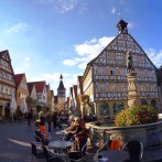 Half-timbered houses in Winnenden with a fountain in the foreground. People sit and walk on the cobbled square under a blue sky., © SMG_Achim Mende