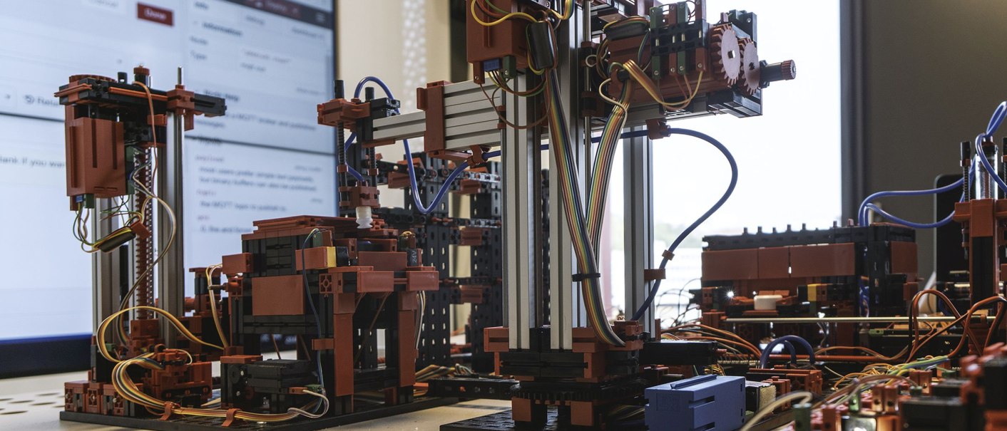 Modell einer IoT-Anlage mit Kabeln und Komponenten, auf einem Tisch vor einem Bildschirm platziert., © INFOMOTION