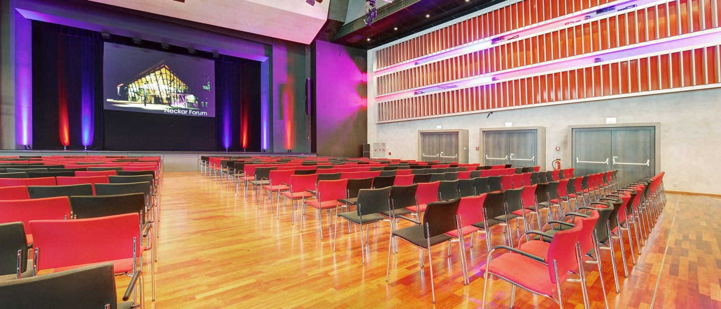 Large hall in the Neckar Forum with empty rows of chairs, stage and colorful lighting. Wooden floor and modern architecture characterize the room., © Martin Frey Large hall in the Neckar Forum with empty rows of chairs, stage and colorful lighting. Wooden floor and modern architecture characterize the room., © Martin Frey