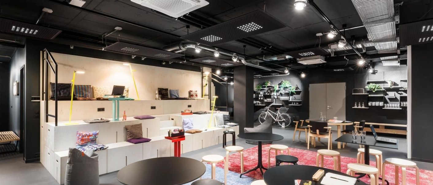 Moderne Lounge mit schwarzen Tischen, hellen Hockern und Regalen. Ein Fahrrad hängt an der Wand, Pflanzen und bunte Teppiche dekorieren den Raum., © Design Offices GmbH