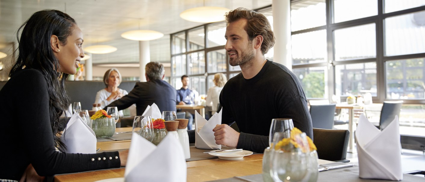Zwei Personen sitzen in einem modernen Restaurant mit großen Fenstern. Der Tisch ist elegant gedeckt mit Gläsern und Servietten., © Mercedes-Benz Kundencenter