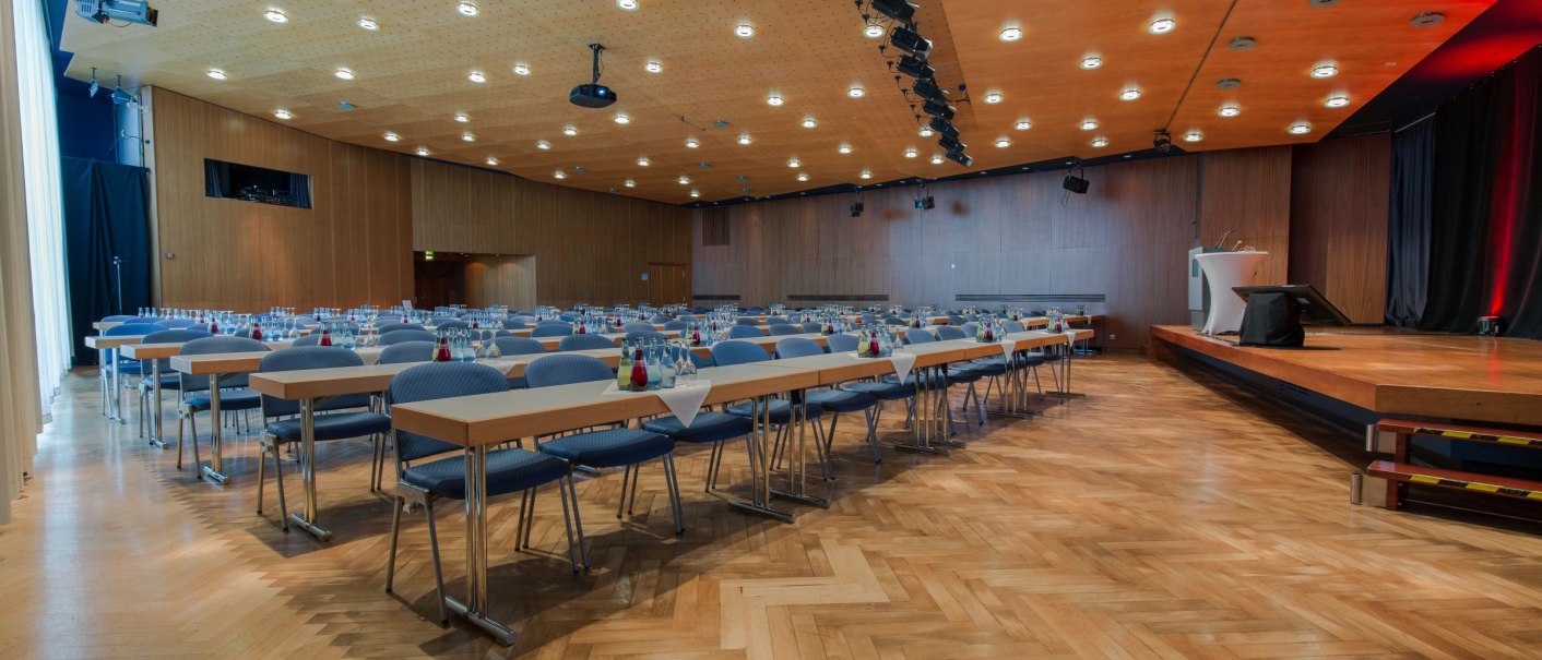 Ein moderner Konferenzsaal mit Holzboden, Tischen und Stühlen, bereit für eine Veranstaltung. Eine Bühne mit Rednerpult ist sichtbar., © ccbs