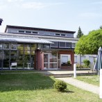 Modernes Gebäude mit Glasfassade, umgeben von Grünflächen und einem Sonnenschirm. Klare Linien und große Fenster prägen die Architektur., © L