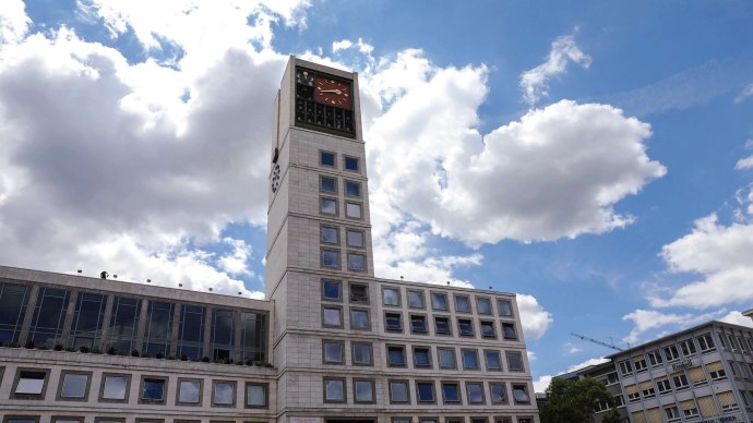 Ein modernes Gebäude mit einem hohen Uhrturm und vielen Fenstern. Der Himmel ist blau mit einigen Wolken., © Fei Shiyu