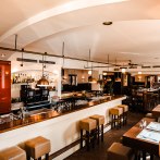 Ein stilvolles Restaurant mit Bar, Holzmöbeln und moderner Beleuchtung. Die Bar ist gut bestückt, und es gibt Sitzgelegenheiten mit gedeckten Tischen., © Amadeus