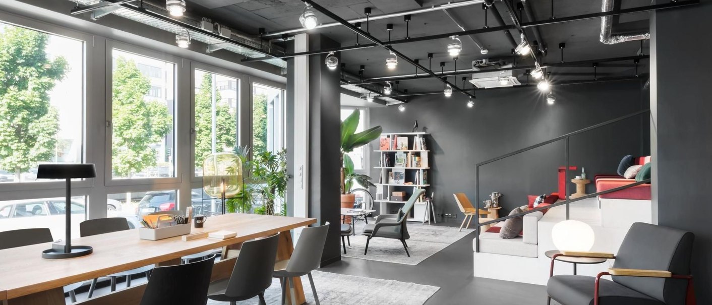 Moderne Lounge mit großem Holztisch, schwarzen Stühlen, Pflanzen und Regalen. Große Fenster lassen viel Licht herein. Gemütliche Sitzecke im Hintergrund., © Design Offices GmbH