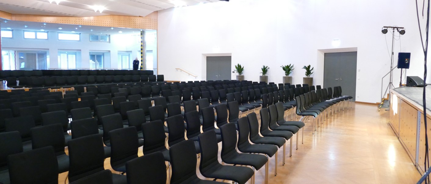 Ein leerer Saal im Kulturzentrum Baltmannsweiler mit schwarzen Stühlen in Reihen vor einer Bühne. Helle Wände und Pflanzen im Hintergrund., © GemeindeBaltmannsweiler_TF Ein leerer Saal im Kulturzentrum Baltmannsweiler mit schwarzen Stühlen in Reihen vor einer Bühne. Helle Wände und Pflanzen im Hintergrund., © GemeindeBaltmannsweiler_TF