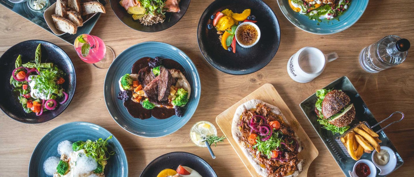 Ein Tisch mit verschiedenen Gerichten: Fleisch mit Gemüse, Salate, Burger mit Pommes, Brot und Desserts. Getränke sind ebenfalls zu sehen., © SatzDrei Ein Tisch mit verschiedenen Gerichten: Fleisch mit Gemüse, Salate, Burger mit Pommes, Brot und Desserts. Getränke sind ebenfalls zu sehen., © SatzDrei