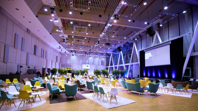 Großer, moderner Raum mit bunten Stühlen und Sofas, Pflanzen und einer großen Leinwand. Die Decke ist mit vielen Lichtern ausgestattet., © Carl Benz Arena