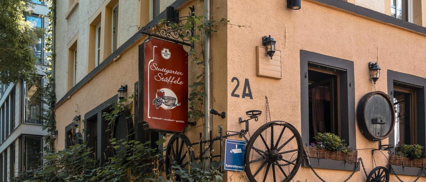Gebäude mit Schild 'Stuttgarter Stäffele', dekoriert mit Pflanzen und alten Wagenrädern. Adresse 2A, Überwachungskamera-Schild sichtbar., © SMG, Sarah Schmid