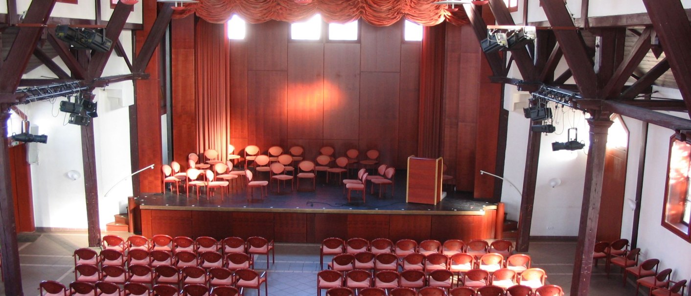 An old gymnasium with wooden beams, a stage and many rows of chairs. The stage is equipped with curtains and a lectern., © Stadt Herrenberg An old gymnasium with wooden beams, a stage and many rows of chairs. The stage is equipped with curtains and a lectern., © Stadt Herrenberg
