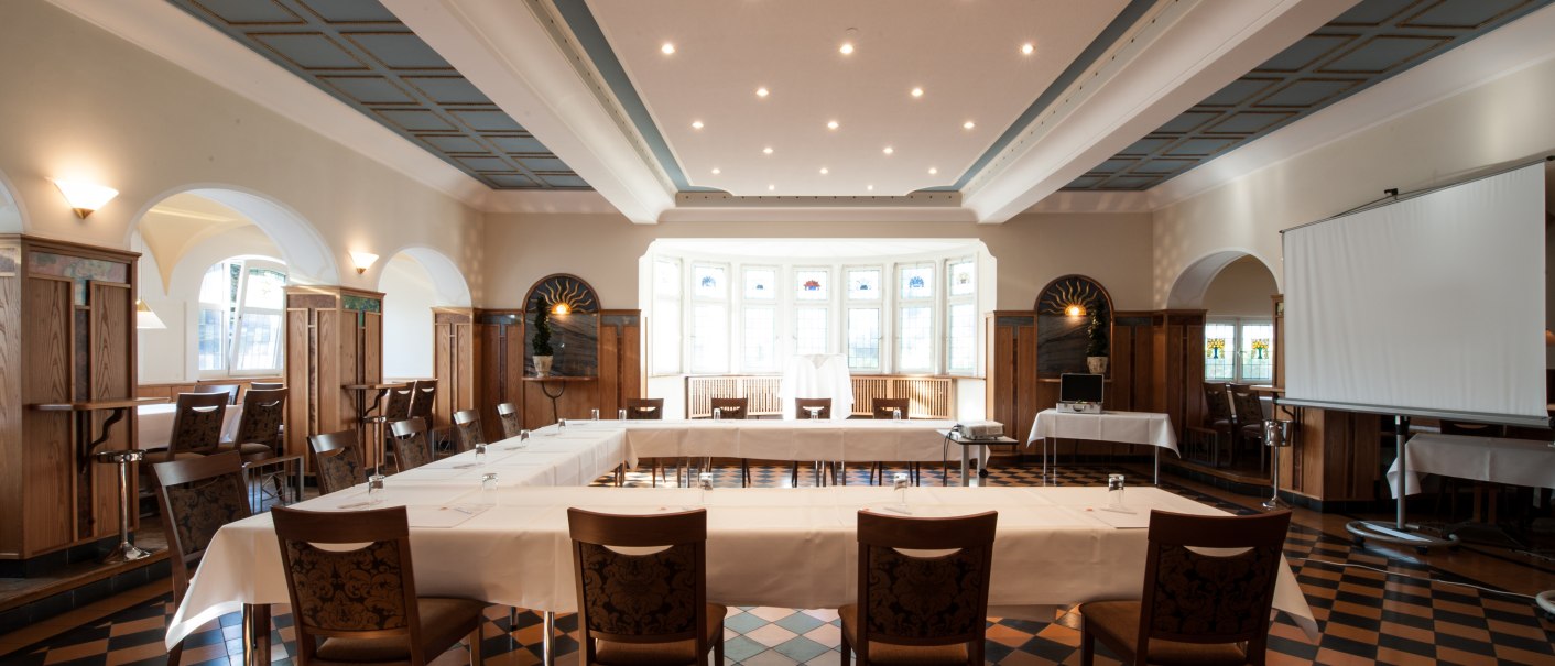 Eleganter Saal mit gedeckten Tischen, Stühlen und einem Projektor. Große Fenster und dekorative Holzelemente prägen den Raum., © Landschloss Korntal Eleganter Saal mit gedeckten Tischen, Stühlen und einem Projektor. Große Fenster und dekorative Holzelemente prägen den Raum., © Landschloss Korntal