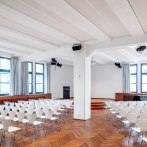 Modern hall with white chairs, wooden floor and stage. Large windows let in plenty of light. Lighting and loudspeakers on the ceiling., © © markus guhl fotografie