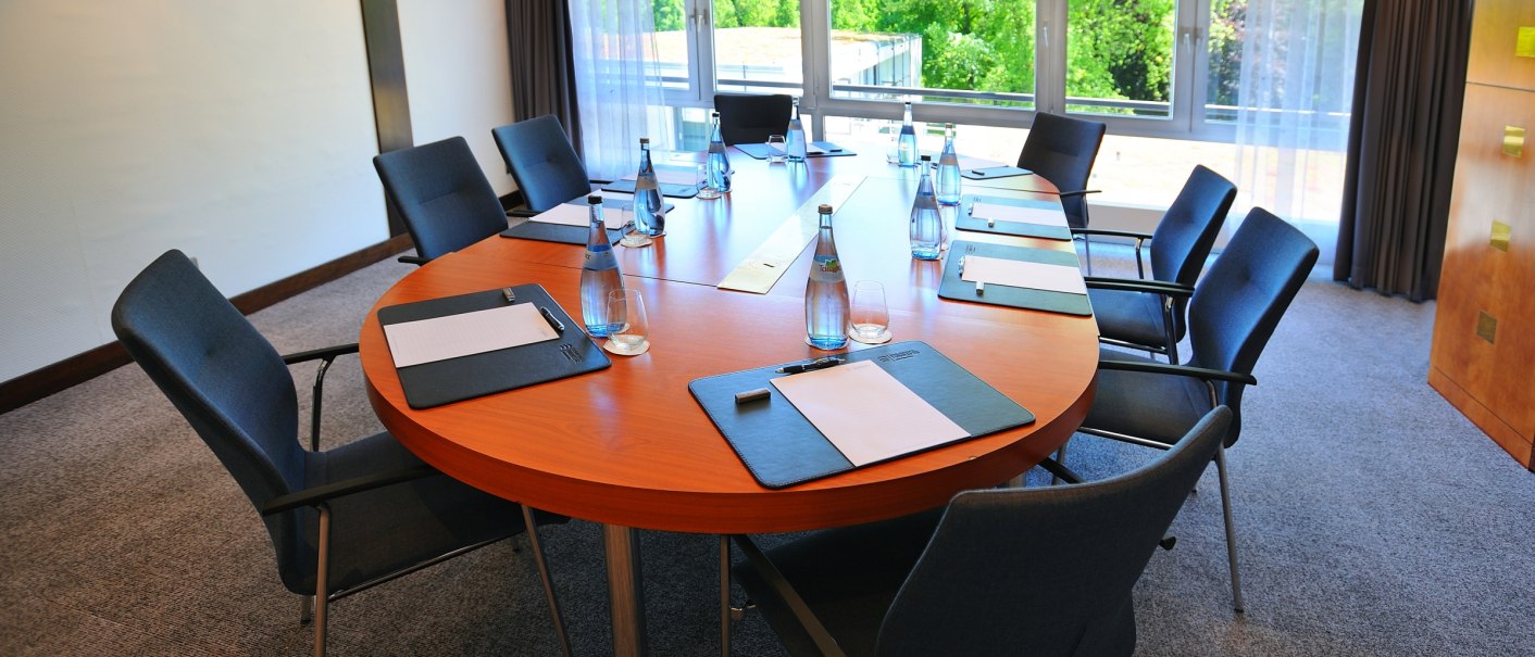 A modern conference room with an oval table surrounded by chairs. There are writing materials and bottles of water on the table. Large windows offer a view of the greenery., © Schlosshotel Monrepos A modern conference room with an oval table surrounded by chairs. There are writing materials and bottles of water on the table. Large windows offer a view of the greenery., © Schlosshotel Monrepos