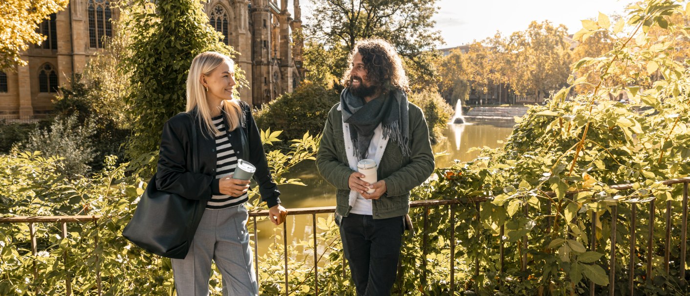 Zwei Personen stehen lächelnd mit Kaffeebechern vor einer Kirche und einem See. Die Szene ist herbstlich und sonnig., © SMG Stuttgart Marketing GmbH - Sarah Schmid