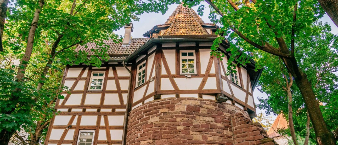 Fachwerkhaus mit rundem Steinturm, umgeben von grünen Bäumen. Das Dach ist mit Ziegeln gedeckt, und die Fenster sind klein und rechteckig., © Thomas Niedermüller Fachwerkhaus mit rundem Steinturm, umgeben von grünen Bäumen. Das Dach ist mit Ziegeln gedeckt, und die Fenster sind klein und rechteckig., © Thomas Niedermüller