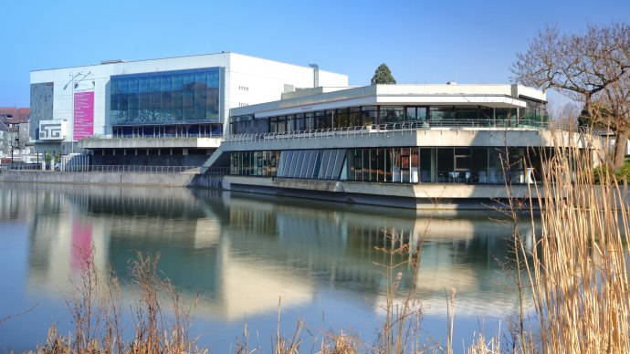Die Kongresshalle Böblingen spiegelt sich im Wasser, umgeben von kahlen Bäumen und Schilf, unter einem klaren blauen Himmel., © ccbs_gf