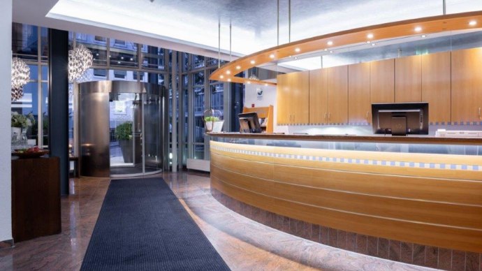 Modern hotel reception with curved wooden counter, illuminated reception area and revolving door in the background. Elegant, inviting ambience., © Kronen Hotel GmbH
