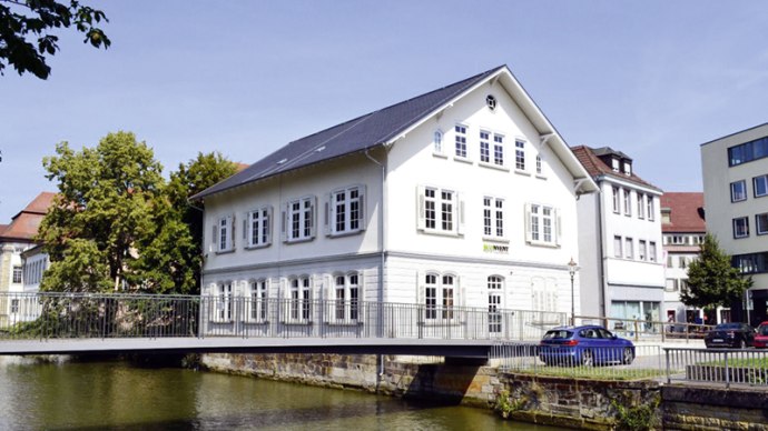 Wei&szlig;es Geb&auml;ude an einem Fluss mit Br&uuml;cke, umgeben von B&auml;umen und weiteren Geb&auml;uden unter blauem Himmel., &copy; econvent