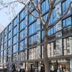 Moderne Glasfassade eines Bürogebäudes in Stuttgart, umgeben von kahlen Bäumen. Menschen gehen auf dem Gehweg entlang. Klare, blaue Himmel im Hintergrund., © Design Offices