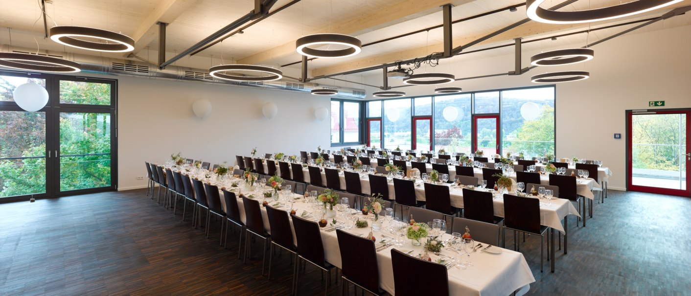 Ein moderner Festsaal mit langen, gedeckten Tischen, großen Fenstern und runden Deckenleuchten. Die Tische sind für ein Event vorbereitet., © Weingaertner-Esslingen-eG Ein moderner Festsaal mit langen, gedeckten Tischen, großen Fenstern und runden Deckenleuchten. Die Tische sind für ein Event vorbereitet., © Weingaertner-Esslingen-eG