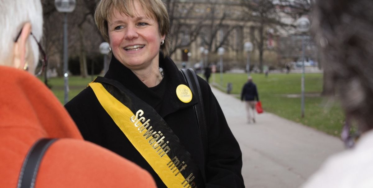 Eine Frau mit einer Schärpe 'Schwäbin mit Herz' lächelt und spricht mit anderen Personen in einem Park., © Stuttgart-Marketing GmbH