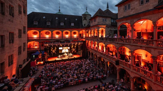 Open-air concert in the courtyard of a historic building, illuminated in red. Spectators sit on balconies and in the courtyard, stage with musicians., © jazzopen stuttgart, Reiner Pfisterer
