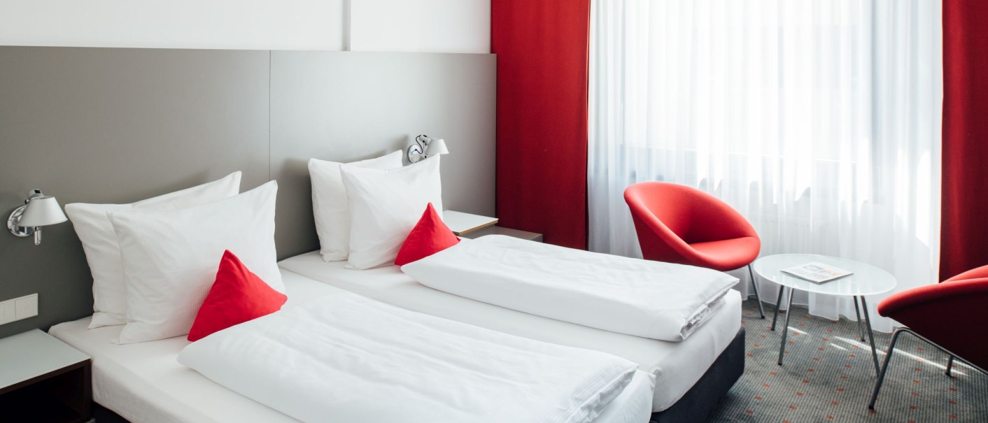 Modernes Hotelzimmer mit zwei Einzelbetten, roten Kissen und Vorhängen. Zwei rote Stühle und ein kleiner Tisch stehen am Fenster., © VHE Mo.Stuttgart
