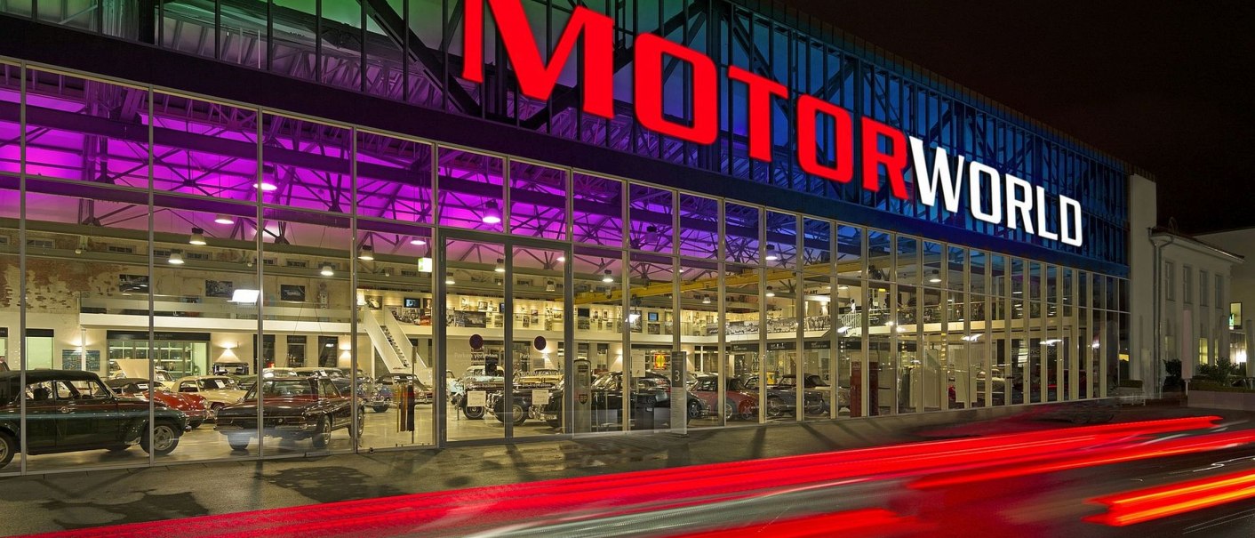 Die Motorworld Region Stuttgart bei Nacht, mit beleuchtetem Schriftzug und bunter Glasfront. Autos fahren vorbei und hinterlassen Lichtspuren., © MW Region Stuttgart Dieter Ege