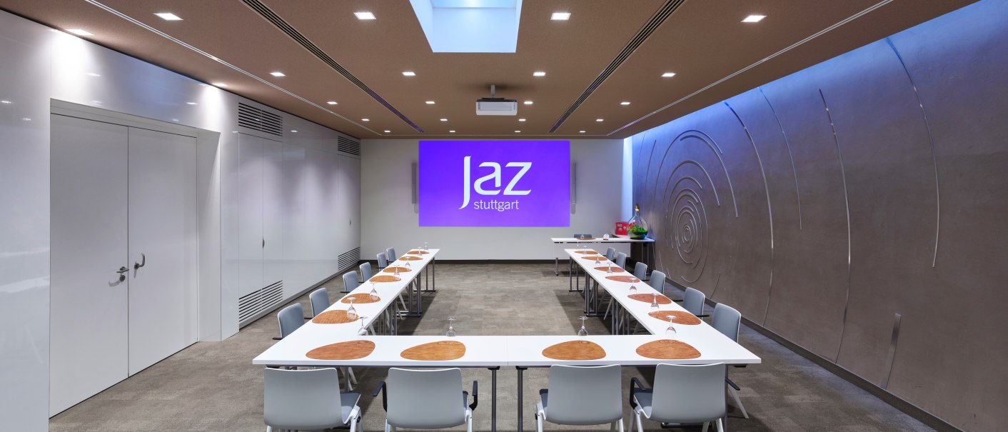 Modern conference room at Jaz in the City Stuttgart with U-shaped table arrangement, chairs and projector. There is a decorative pattern on the wall., &copy; JazHotels