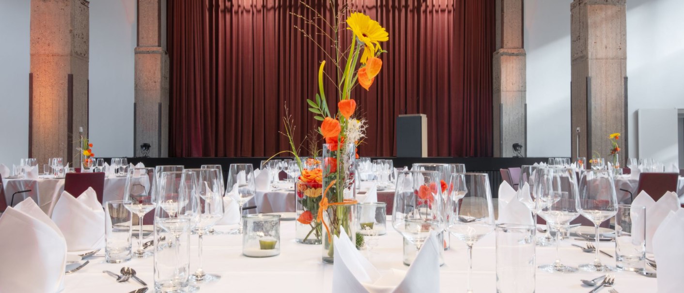 Elegant ballroom with tables set in white, floral arrangements and a stage with a red curtain in the background., © Stefan Baraniecki Elegant ballroom with tables set in white, floral arrangements and a stage with a red curtain in the background., © Stefan Baraniecki